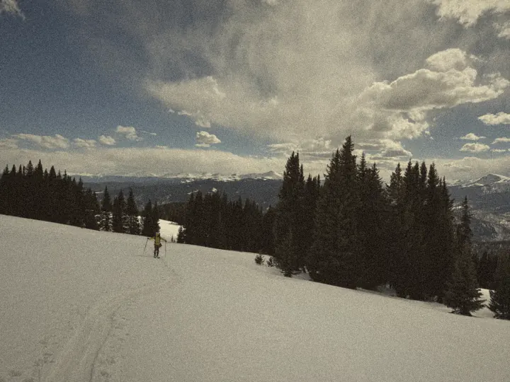 A skier in a yellow jacket climbing a skin track through a snowy meadow, peaks on the horizon