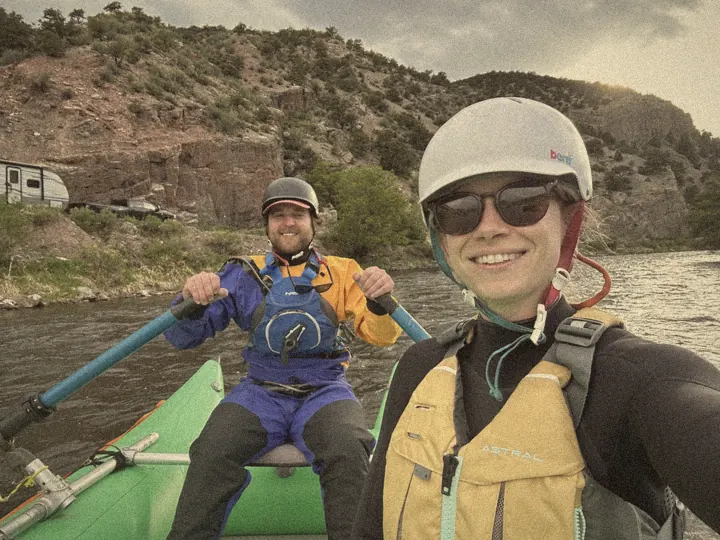 Selfie on a green raft, one person rowing in a drysuit, red canyon walls behind
